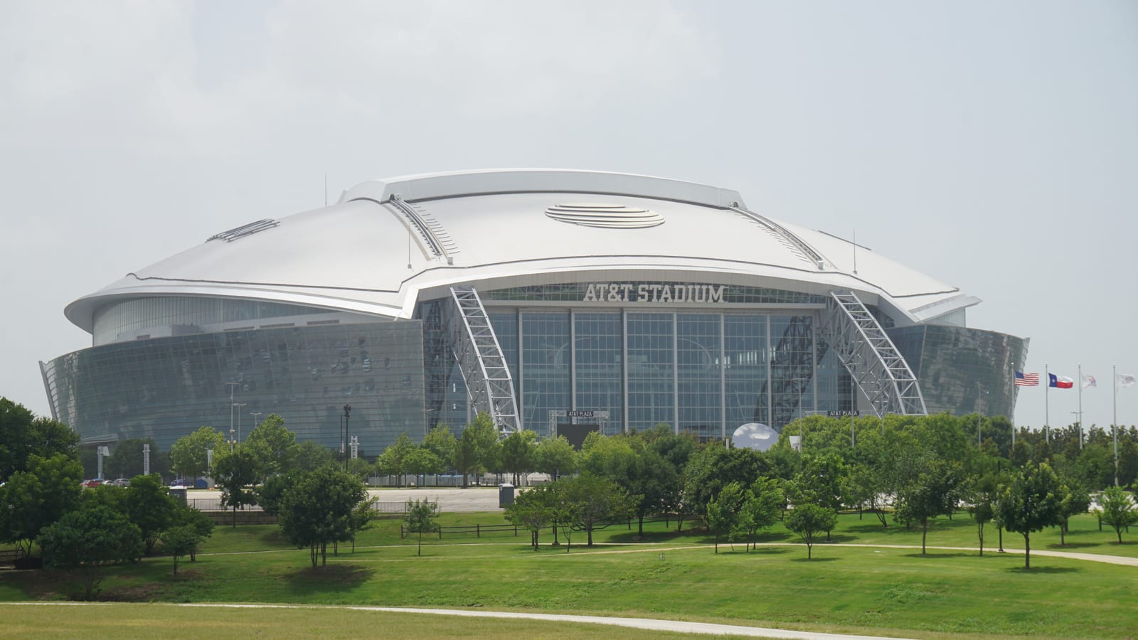 A domed stadium with a glass facade and arched steel supports stands behind a wide lawn and a row of trees.