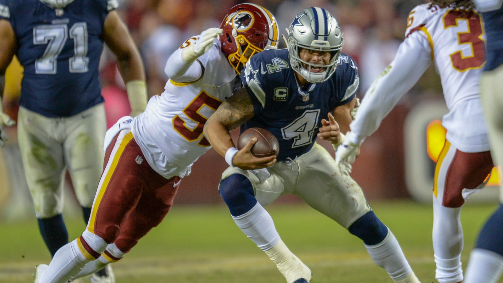 A quarterback in a navy number 4 jersey and silver helmet carries the ball as a defender in white reaches for him.
