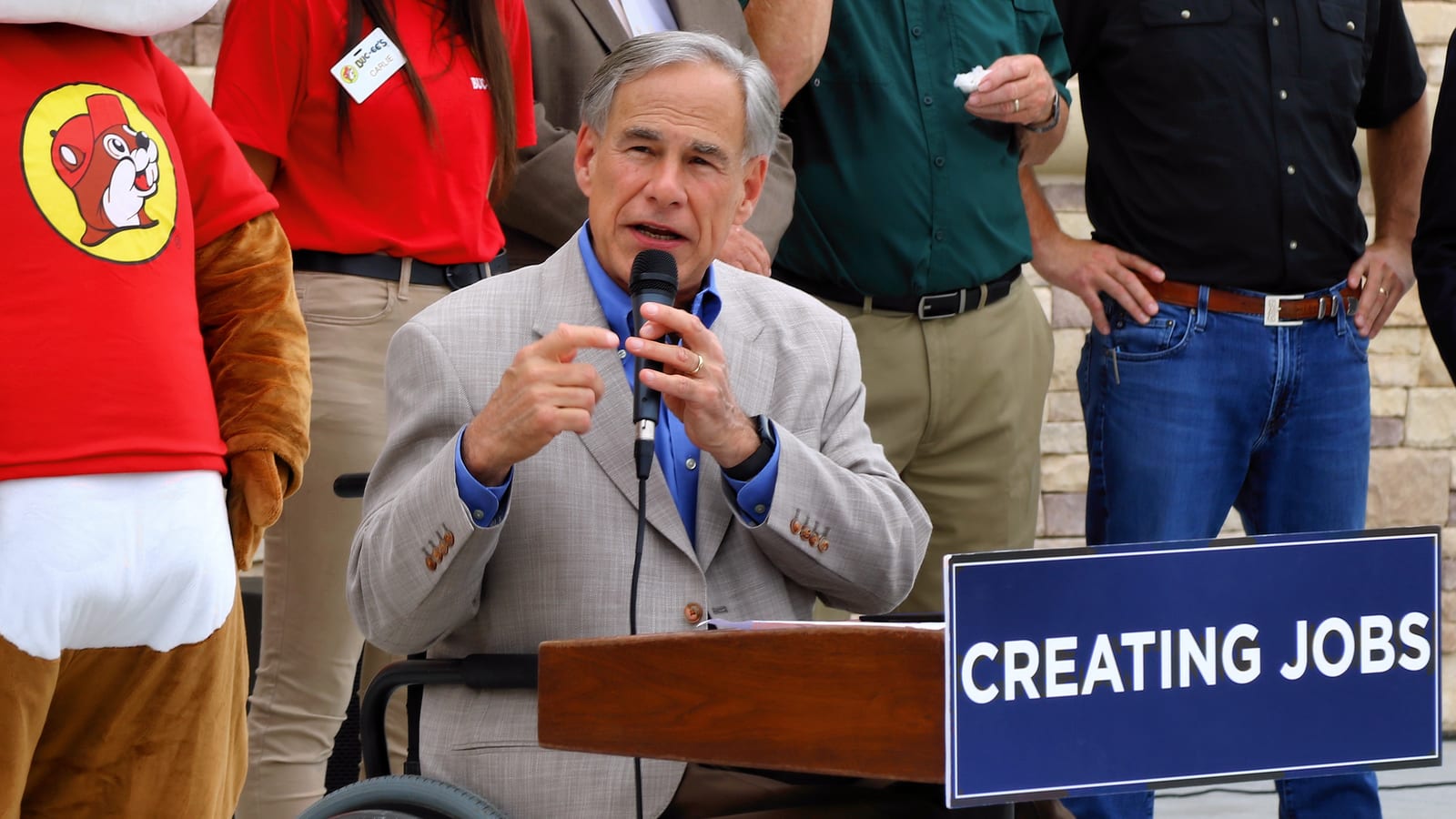 A gray-haired man in a tan sport coat speaks into a handheld microphone at a lectern beside a sign reading Creating Jobs, with people and a beaver mascot standing behind him.