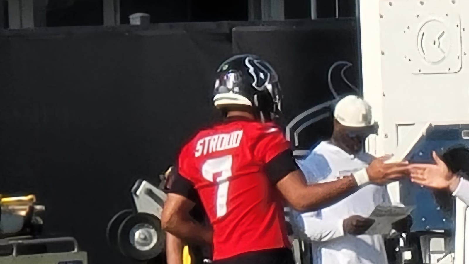 A quarterback in a red practice jersey with the name Stroud and the number 7 walks along the sideline at a practice field, slapping hands with a staff member.