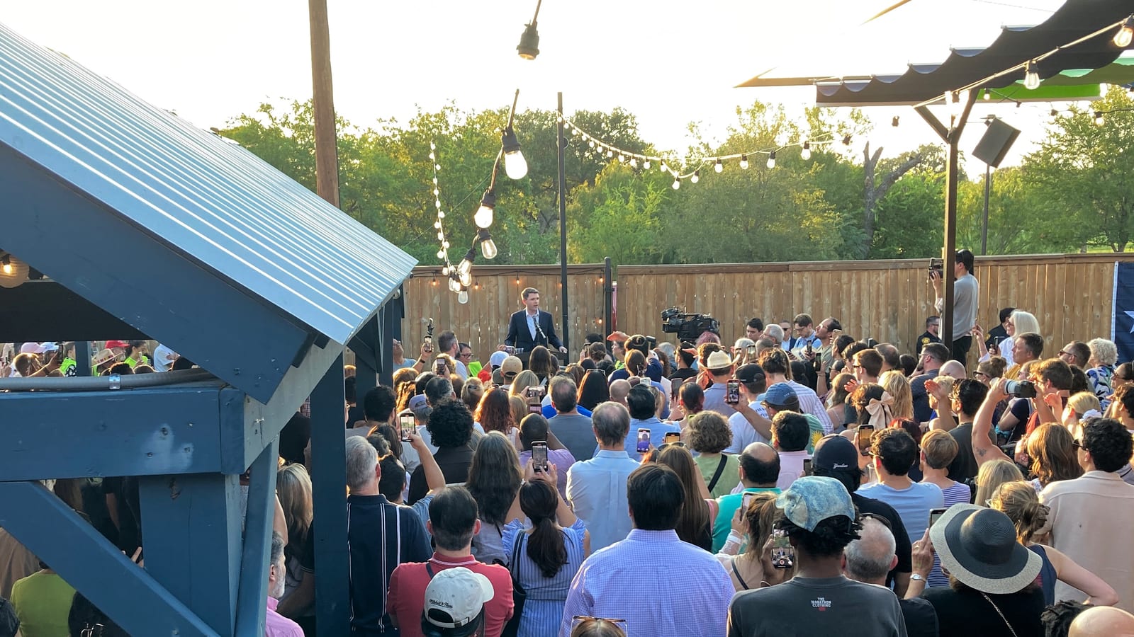 A man in a dark suit speaks from a small stage under string lights to a large outdoor crowd, with a Texas flag on the fence behind the audience.