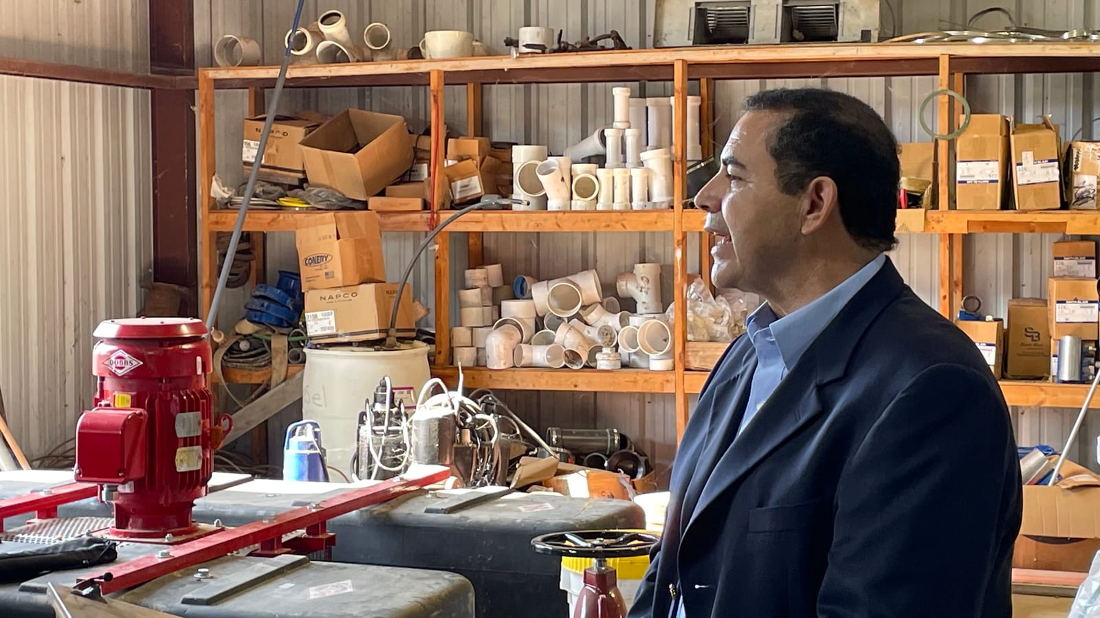 A man in a navy blazer and light blue shirt stands in profile in a metal workshop lined with shelves of white plastic pipe fittings, a red pump motor in the foreground.