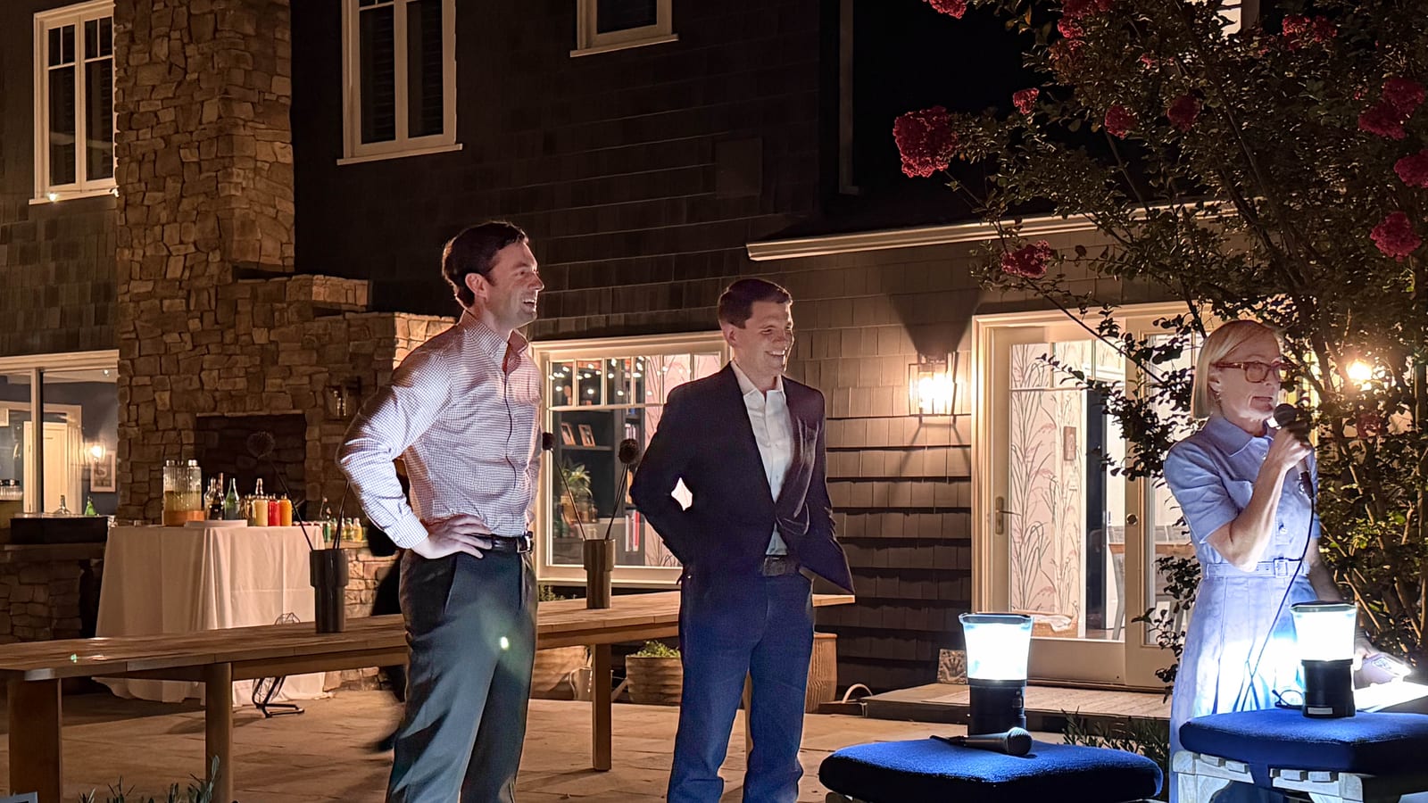 Two men stand on a lit patio at night outside a shingled house, one in a checked shirt and gray trousers and the other in a navy blazer and jeans, while a woman at right speaks into a microphone.