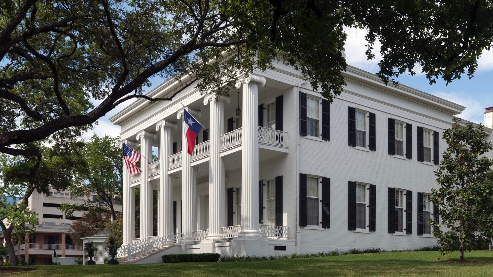 A white two-story Greek Revival house with tall fluted columns, black shutters and a United States flag and a Texas flag on the porch, framed by oak branches under a partly cloudy sky.